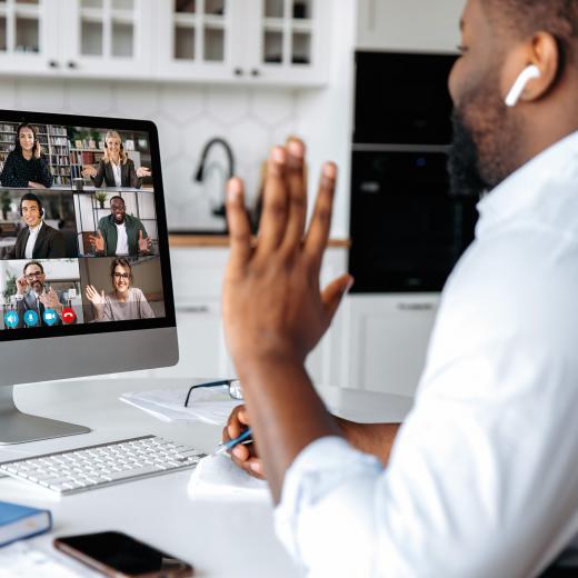 A person sitting at a desk waving to a monitor showing several people on a conference call