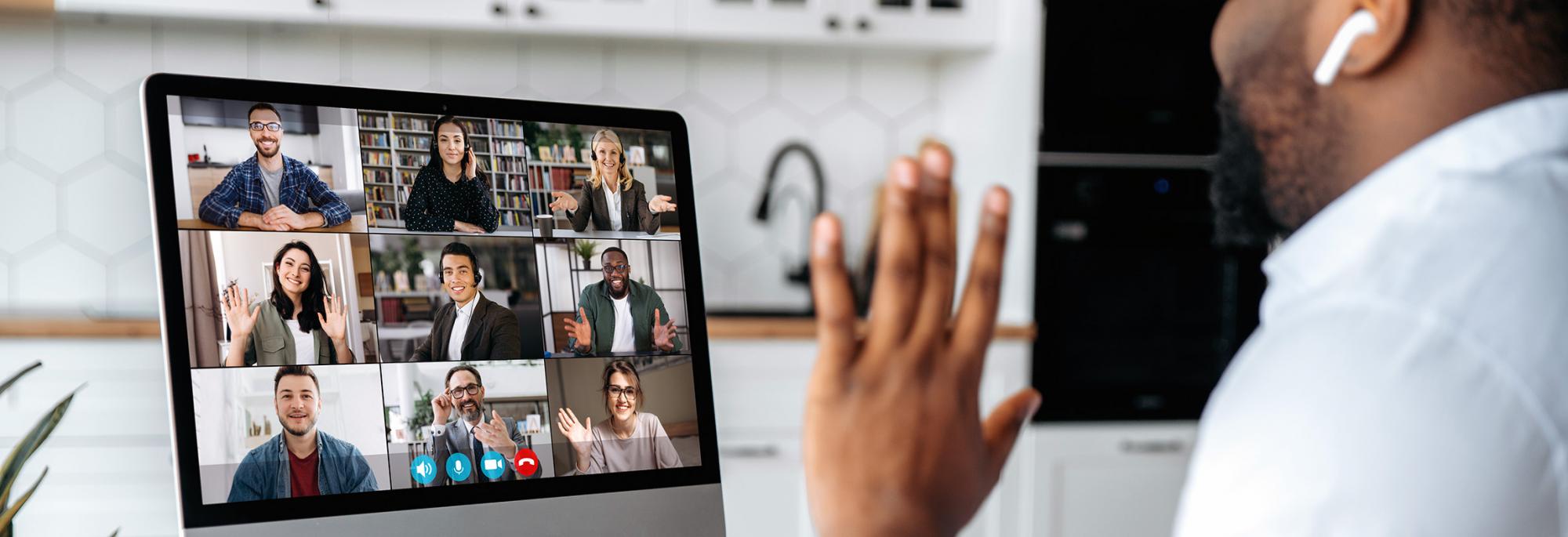 A person sitting at a desk waving to a monitor showing several people on a conference call