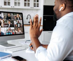 A person sitting at a desk waving to a monitor showing several people on a conference call